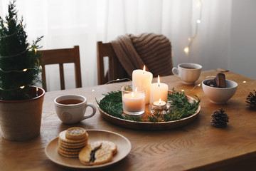 Fototapeta na wymiar Cozy evening tea party by candlelight. Served table: cup of tea, cookies, tray, candles, pine cone, branches, little christmas tree in a pot, led garland lights. Table setting with holiday decorations
