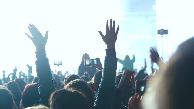 Crowd of spectators raises their hands up and applauds the musician performing on stage. Spectators at the concert photographed artist. Big crowd at concert cheering clapping hands at night concert.