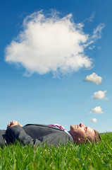 Businessman lying down outdoors in bright green grass dreaming under thought bubble clouds in blue skies