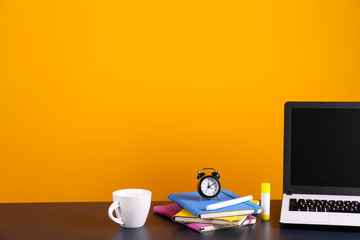 Blank screen laptop computer, various supplies on wooden desk over bright yellow wall background with a lot of copy space for text. Creative workspace. Close up shot of table w/ computer & stationery.