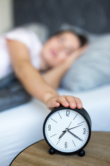 morning concept - young sleepy woman awaking up with alarm clock in bedroom
