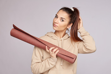 Close-up tender and feminine young mixed race woman with dark hairstyle, playing with pony tail gazing camera, smiling charming as standing grey background, holding fitness mat, wearing casual hoodie