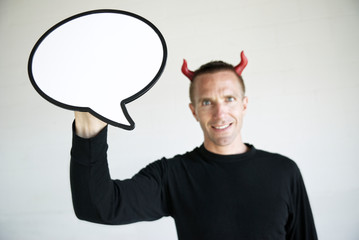 Smiling man with red devil horns holding a large cartoonish speech bubble looking at the camera with a mischievous expression