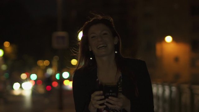 Caucasian modern woman wearing flower dress, black jacket and red hair walking through the street and writing a text message on smartphone and laughing at camera by night. Paris 4K UHD. Slow Motion.