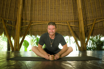 attractive happy man in hipster yogi style doing yoga drill sitting on mat at beautiful Asian bamboo hut stretching body enjoying nature in mind balance and good vibes