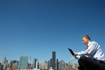 Businessman sitting outdoors using tablet computer at the city skyline under clear blue sky copy space