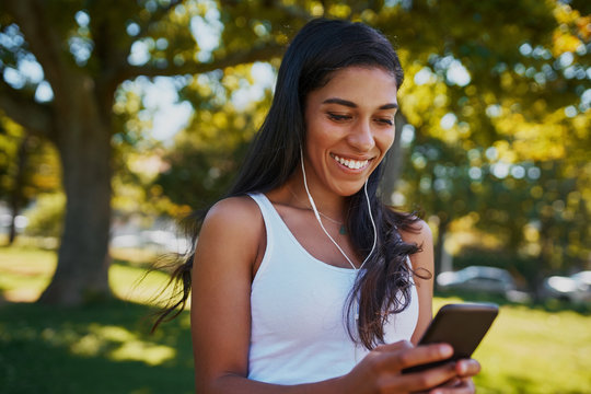 Portrait of a smiling young woman with earphones in her ears using mobile phone to listen to music in the park under a tree on a sunny day