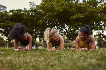 Three sporty young women smiling while doing plank exercises lying on green grass in the park