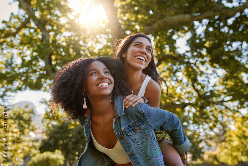 Poster two diverse multiracial friends having fun laughing and giving a piggyback to each other in the park