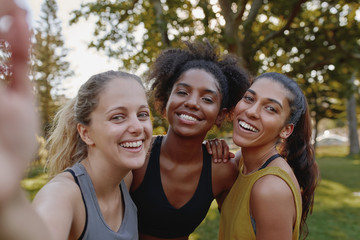 Smiling portrait of diverse female friends looking at camera taking selfie in park - fitness friends taking a selfie after exercising 