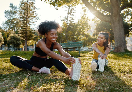 Young diverse female friends sitting on green grass stretching her legs in the morning sunlight at park - diverse friends warming up before doing group exercise 