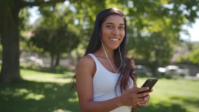 Cheerful young brunette mixed race woman with earphones in her ears look at camera and smiles white texting messages on mobile phone in the park
