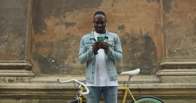 Handsome Afro American Man in Earphones Wearing Jean Jacket Using his Modern Smartphone. He Standing in front of Modern Bike and Old Building Wall at the Background Close Up.