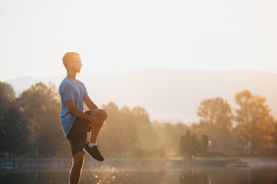 Young Man In Sportswear Stretching By The Lake In A City Park. Runner Warming Up In The Park By The Water In The Early Morning.