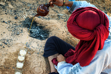 Moroccan man with red turban prepares traditional mint tea in the desert.