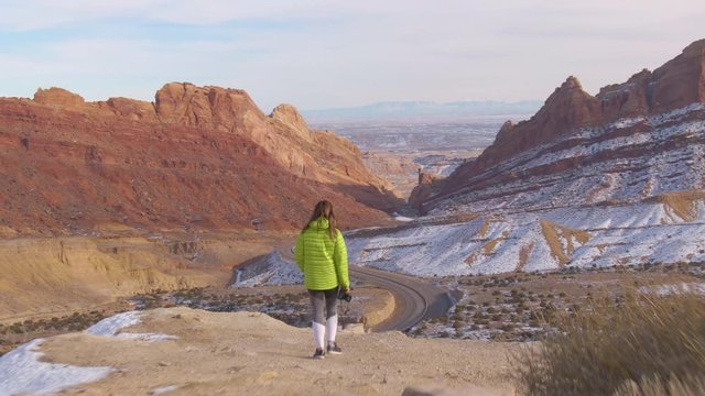 DRONE: Female traveler walks with her camera in hand along a scenic trail overlooking an asphalt highway crossing the canyon. Instagrammer walking to a good photo spot in a stunning geological park.