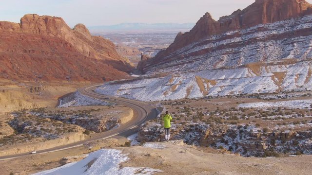 AERIAL: Flying around a woman taking photos of highway crossing the snowy canyon from edge of a cliff. Stunning drone shot of female trekker taking pictures of sandstone formations overlooking a road.