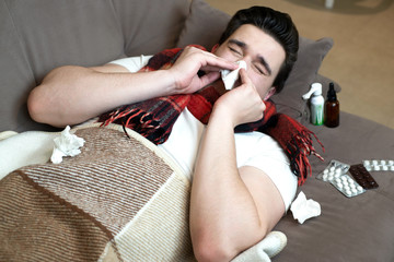 young man lying on the sofa with fever feeling sick bowing his nose with paper handkerchief surrounded with pills and spray illness at home