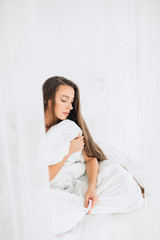 A young woman sitting on bed wrapped in a blanket. Morning in white room.