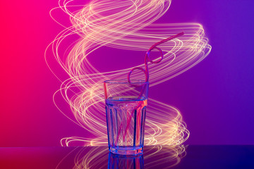 Glass with beverage, curly straw in neon pink and blue light on mirror bar with light painting. Night club, party, Birthday concept. Close-up, copy space