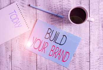 Writing note showing Build Your Brand. Business concept for enhancing brand equity using advertising campaigns Stationary placed next to a cup of black coffee above the wooden table