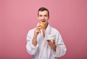 Perky cool guy in white bathrobe is standing on wall back with fragrant croissant