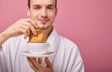 Funny guy in snow-white bathrobe is dipping croissant in tea