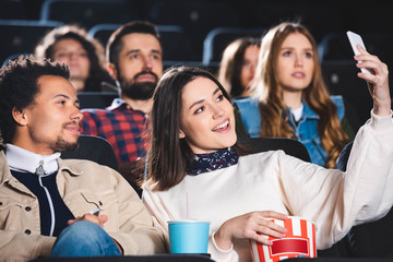 selective focus of multiethnic friends taking selfie and smiling in cinema