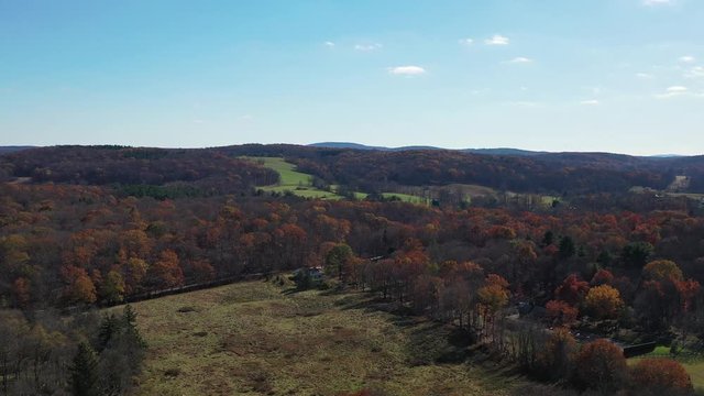 aerial dolly in towards the horizon, over fall foliage tree tops, moving slowly towards a green pasture in the distance. It is a sunny day in bucolic Millbrook, NY