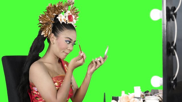 Attractive balinese dancer applying makeup on her face while holding a small mirror in the studio. Shot in 4k resolution with green screen background