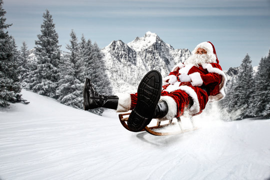 Red Santa Claus Riding A Wooden Sled. An Older Man With A Beard Delivers Presents To A Child. Winter Mountains Landscape And Snow-covered Trees With Frost. Fir Branches, Winter Time. Christmas Spells