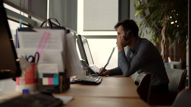 Creative Man With Computer Working At Open Space Room In Sunset.Overtime Work In Office.Exhausted Businessman. Businessman Talking On Phone In Modern Office.Man Working In Office And Typing Keyboard.