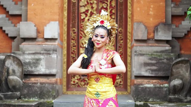 Beautiful balinese dancer smiling at the camera while showing a welcoming gesture in traditional costume at temple. Shot in 4k resolution