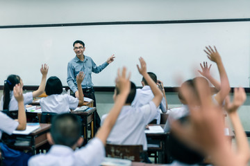 An smiling Asian male high school teacher teaches the white uniform students in the classroom by asking questions and then the students raise their hands for answers.