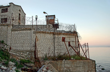 Old fortified prison wall adjacent to the sea. Barbed wire on the walls. Security tower. Calm sea.