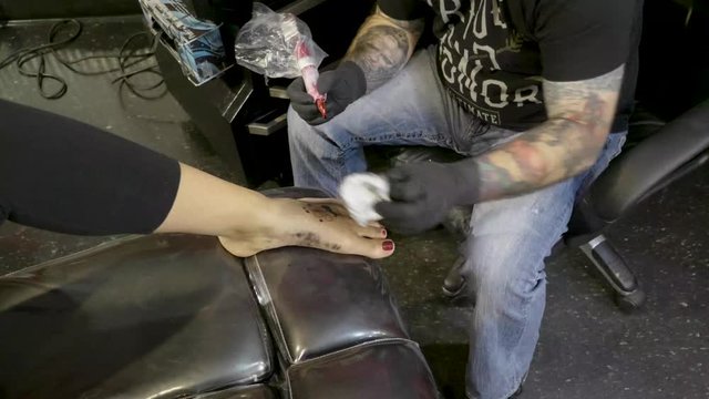 Tattoo artist placing small black and white tattoo on ladies foot wide shot, POV