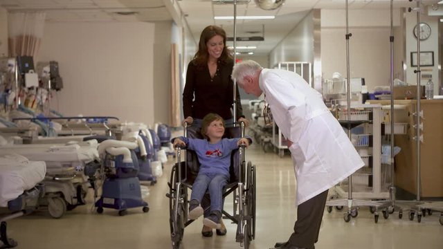 A friendly doctor talks to a mother and her son in the hospital as she wheels her son up in a wheelchair.
