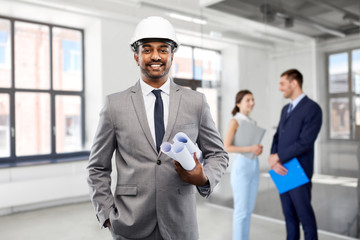 architecture, construction business and occupation concept - smiling indian male architect in helmet with blueprints over people in empty office room background