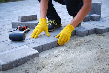 The master in yellow gloves lays paving stones in layers. Garden brick pathway paving by professional paver worker. Laying gray concrete paving slabs in house courtyard on sand foundation base.