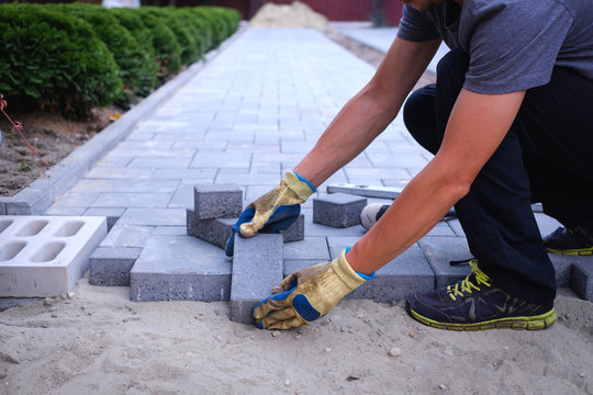 The master in yellow gloves lays paving stones in layers. Garden brick pathway paving by professional paver worker. Laying gray concrete paving slabs in house courtyard on sand foundation base.
