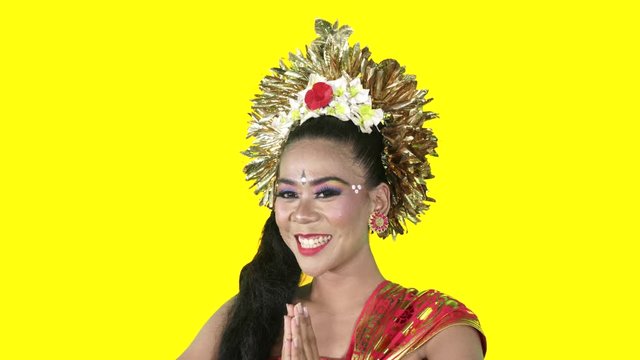Friendly balinese dancer in traditional costume showing a welcoming gesture while smiling at the camera in the studio. Shot in 4k resolution with yellow background