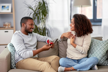 anniversary, proposal and couple concept - happy african american man giving diamond engagement ring in little red box to woman at home