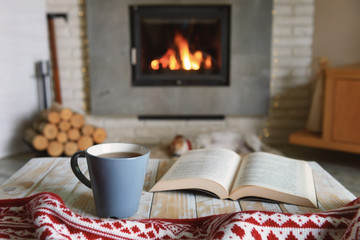 Hygge concept with open book and cup of tea near burning fireplace