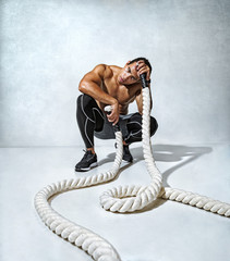 Man taking a break from workout sitting on her toes holding battle ropes. Photo of sporty man relaxing during workout on grey background. Strength and motivation. Full length