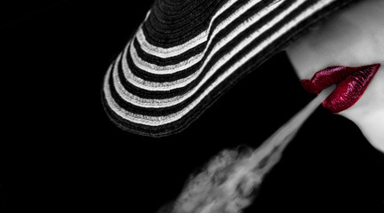 Annonymous beautifaul artistic woman with full red lips with stripped hat hiding eyes blowing smoke out of her mouth with copy space on black backround.
