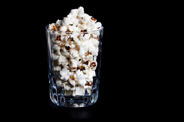 many crunchy popcorns are in clear glass on dark black background, food or sneak for the movies and the circus. A dark high angle photo. Traditional food is prepared, horizontal orientation.