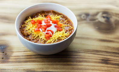 Bowl of red chili with tomatoes, sour cream and cheese toppings. Wood background, copy space. 