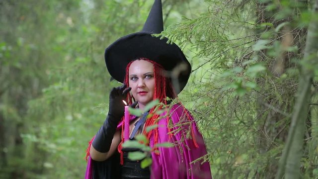 a young witch with red hair and a pointed hat and a black cloak conjures with candles in the forest.