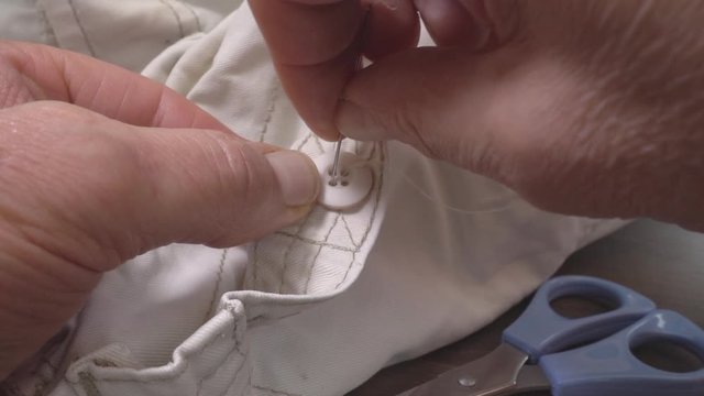 Slow motion close POV shot of a man&rsquo;s hands sewing a button onto a pair of cotton trousers.