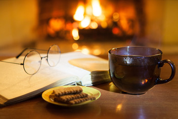 An open ancient book, vintage glasses, a cup and biscuits in front of the fireplace. Evening fairy tales. Fantasy.